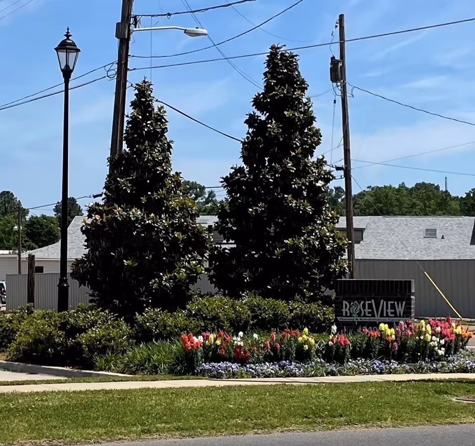 Outdoor view of the entrance area of Roseview Nursing & Rehabilitation Center featuring a landscaped garden with colorful flowers, two tall evergreen trees, a street lamp, utility poles with wires, and a sign displaying the facility's name.