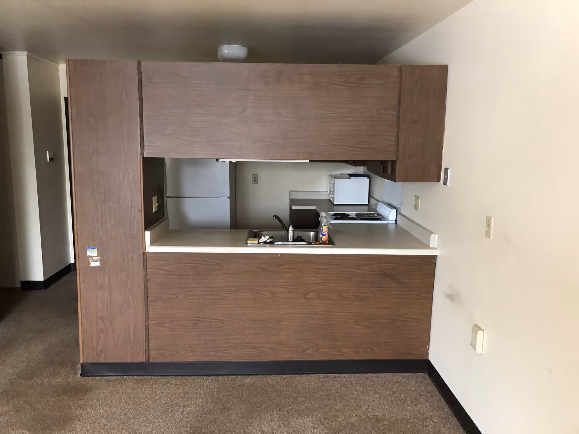 View of a small kitchen area with brown wooden cabinets, a white countertop, a sink, a white refrigerator, a microwave, and a stove. The kitchen is partially enclosed by a wooden partition with an open window-like space. The surrounding walls are beige, and the floor is carpeted.