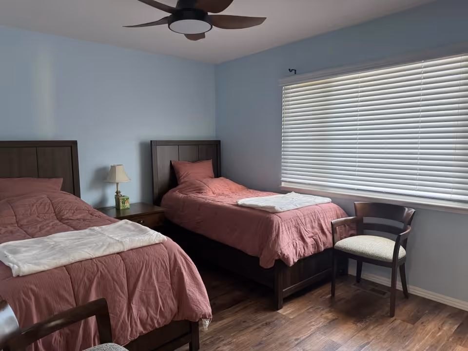 A bedroom with two single beds featuring dark wooden headboards and pink bedding. Each bed has a white folded blanket at the foot. Between the beds is a wooden nightstand with a small lamp and a decorative box. A wooden chair with a cushioned seat is positioned near a large window with closed white blinds. The room has light blue walls, a wooden floor, and a modern ceiling fan with dark blades.