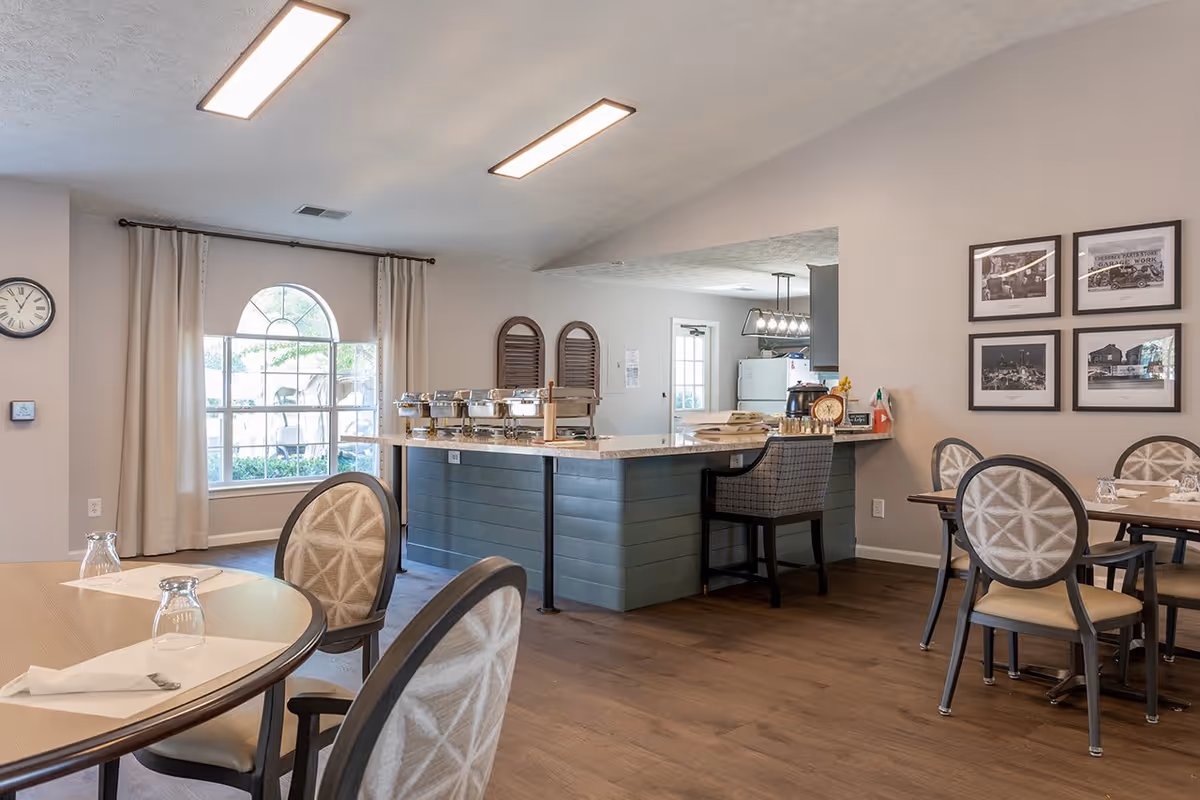 A dining area in a senior living facility featuring round tables with chairs, place settings with napkins and glasses, a large window with curtains, and a kitchen counter with buffet-style food warmers. The room has wooden flooring, framed black and white photos on the wall, and bright overhead lighting.