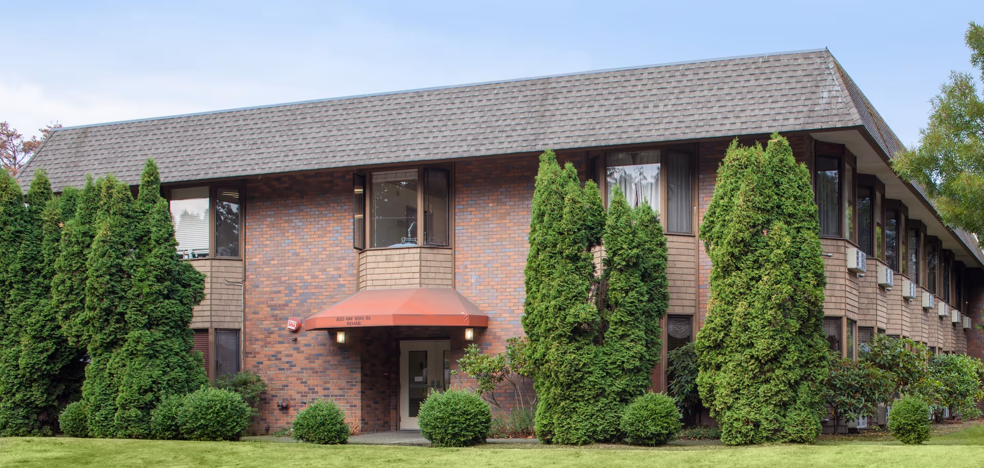 Front exterior of a two-story brick building with a red awning over the entrance, surrounded by tall evergreen shrubs and a manicured lawn.