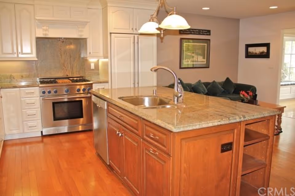 Bright kitchen with island, stainless steel stove, white cabinets, and living room visible in background.
