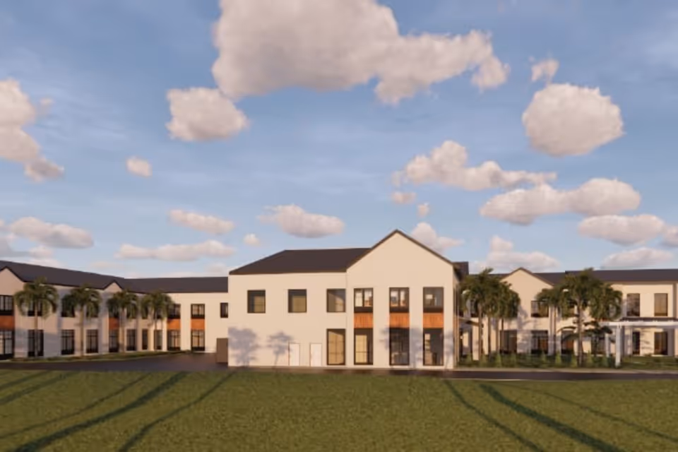 Exterior view of a two-story building with white walls and dark roof, surrounded by palm trees and green lawn under a partly cloudy sky.