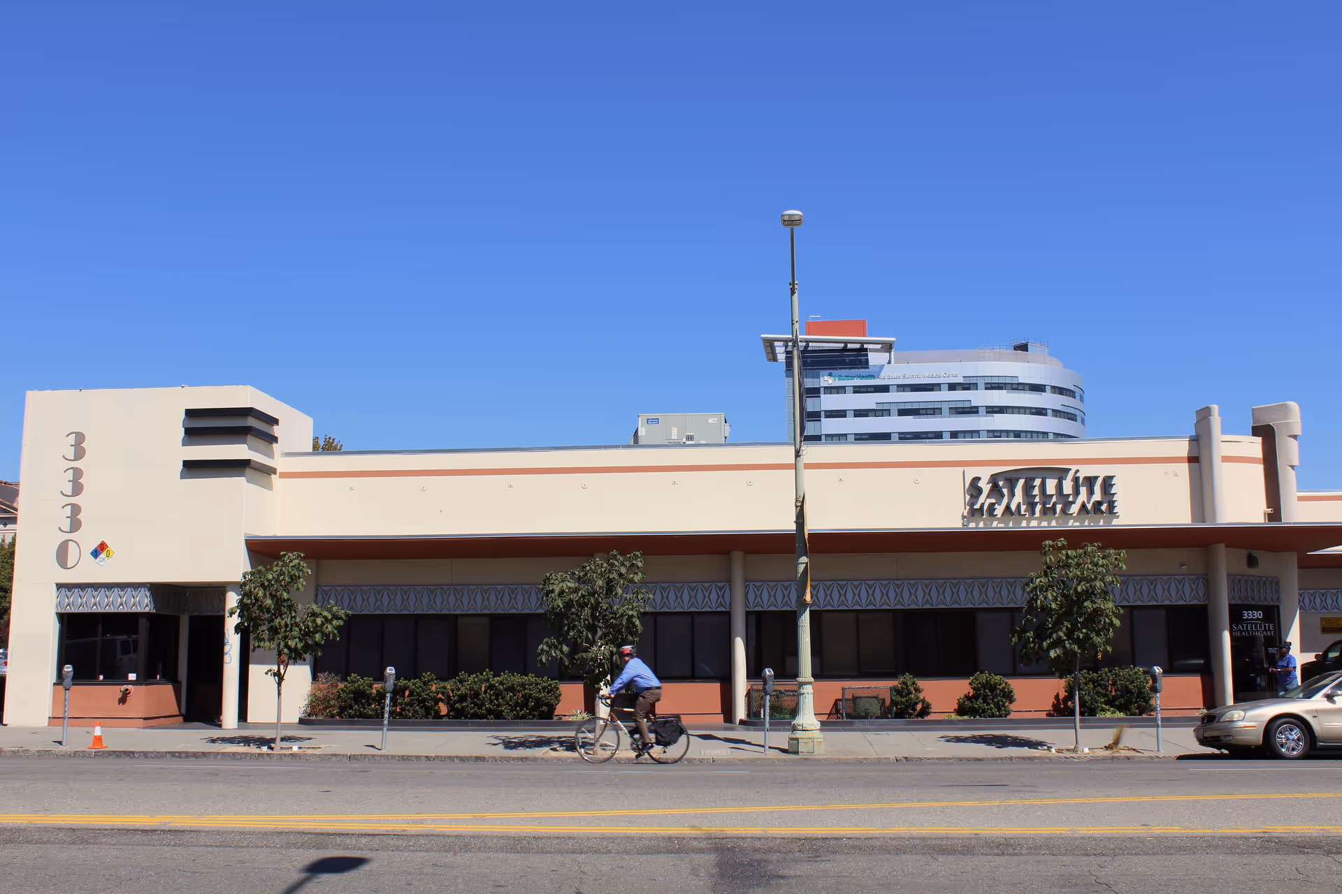 Sunlit exterior of Satellite Healthcare Oakland building with street, cyclist, and parked cars.