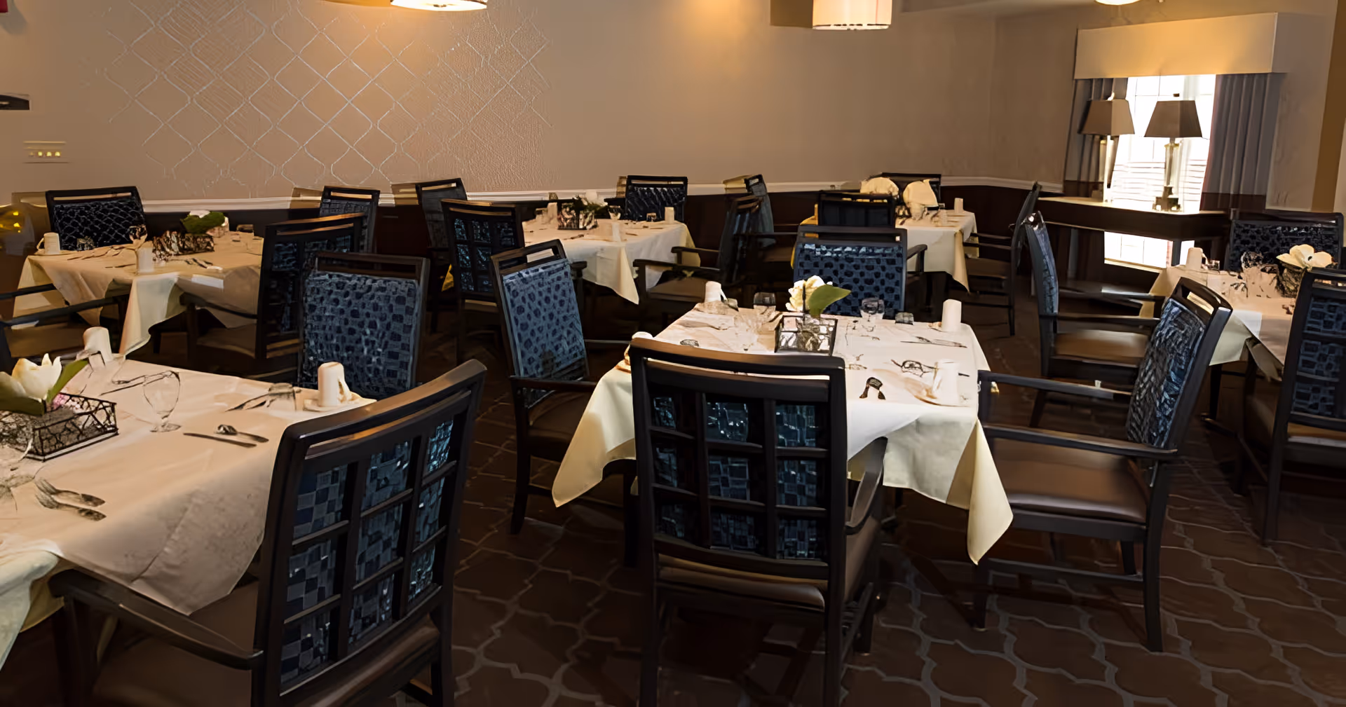 Dining room with multiple tables covered in white tablecloths, set with glassware, napkins, and silverware. The chairs have dark wood frames and blue patterned upholstery. The room has soft lighting, a window with curtains, and two table lamps on a sideboard.