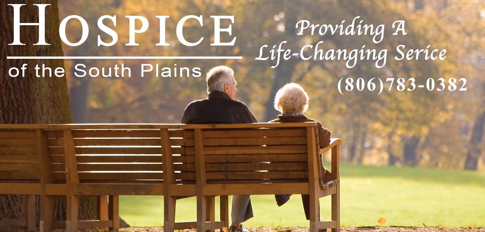 Two elderly individuals sit on a wooden bench in a sunlit park.