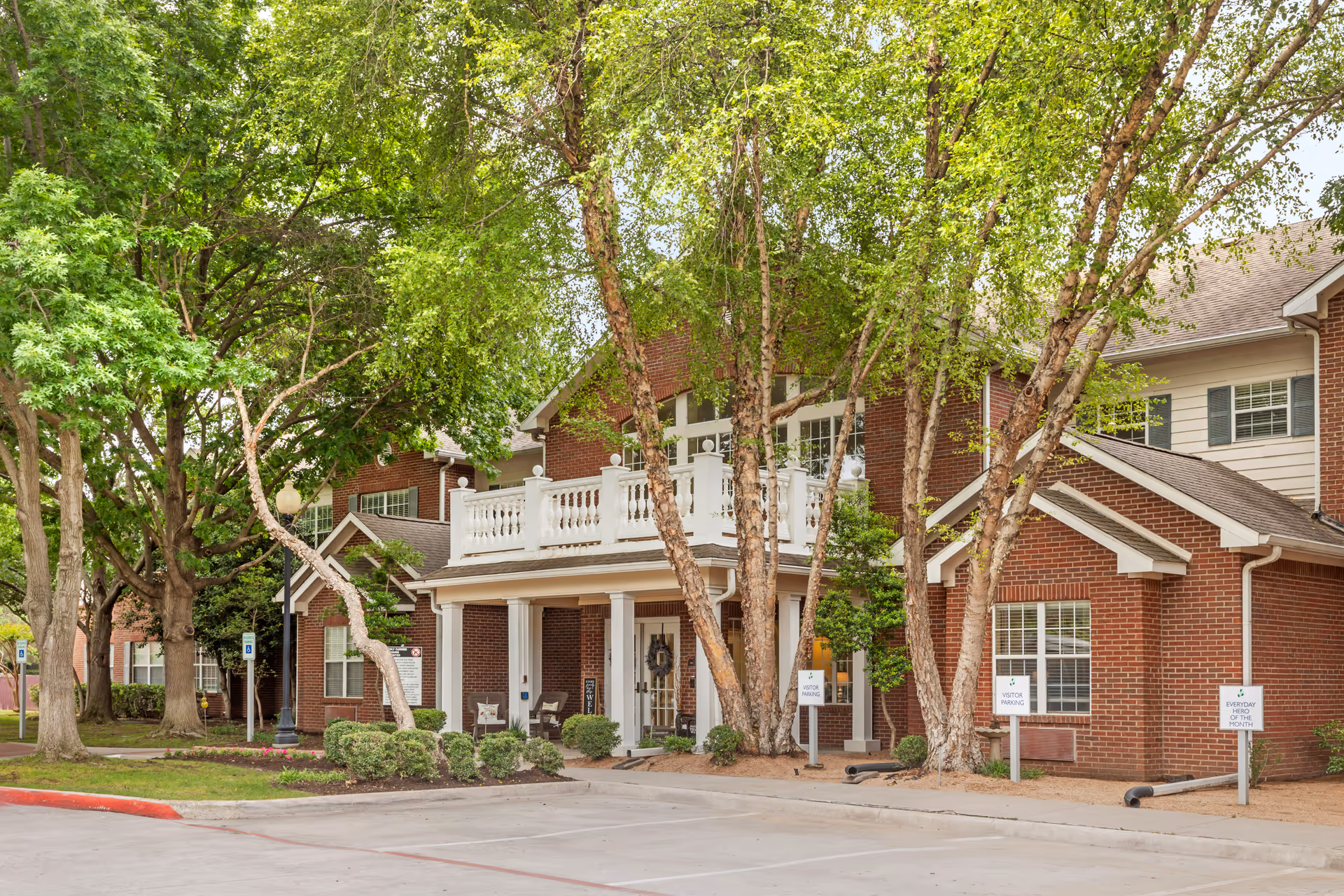 Brick senior living facility entrance with columns, a second-floor balcony, and mature trees.