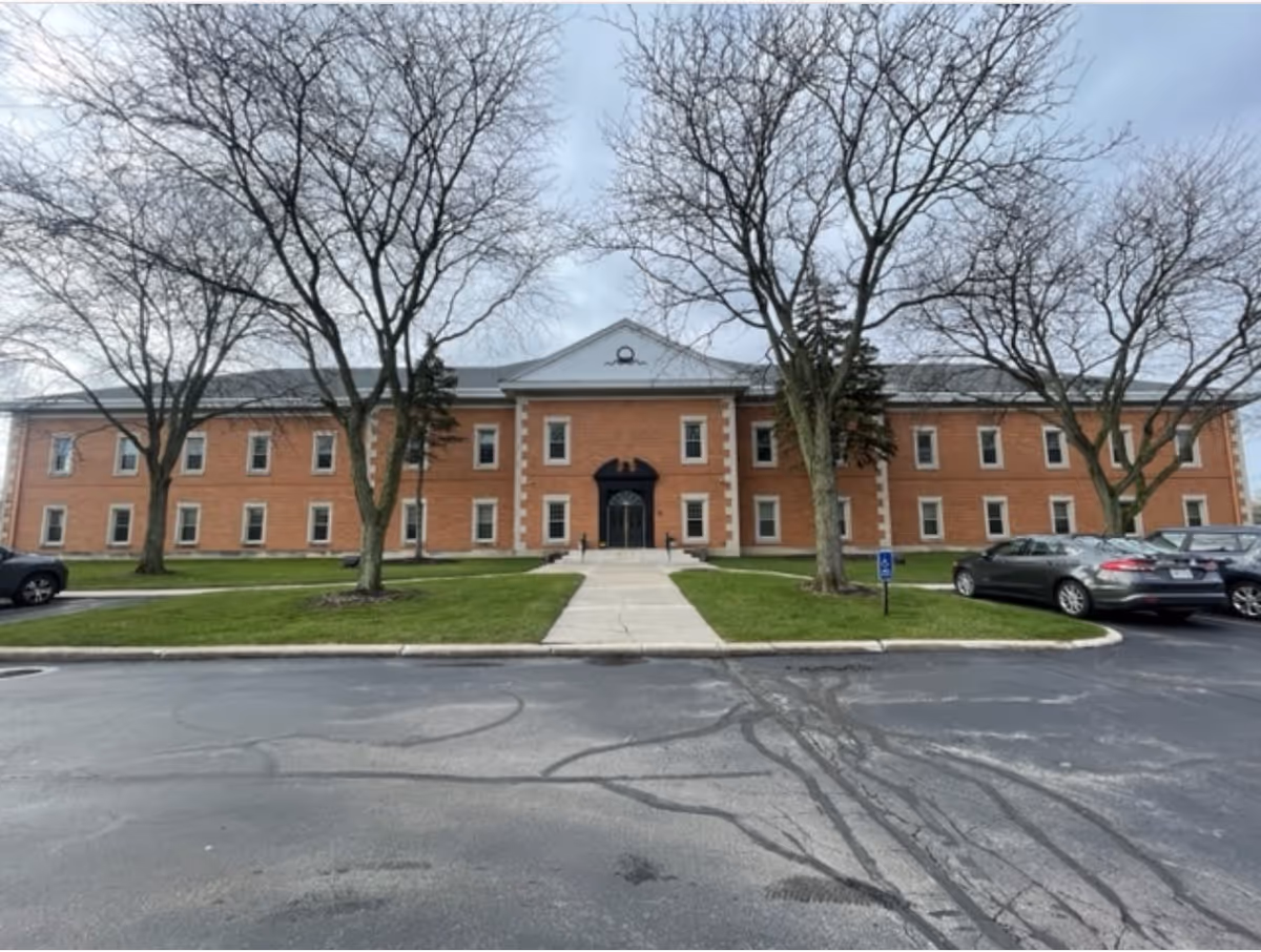 Brick senior living facility facade with trees, a central entrance, and parked cars.