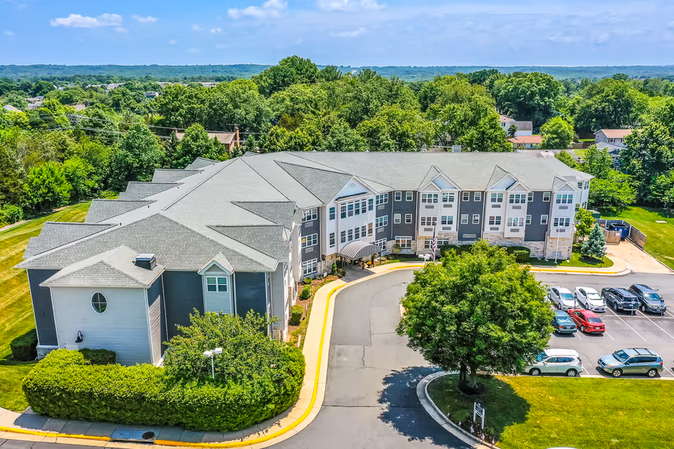 Aerial view of a large senior living facility building with gray and white exterior, surrounded by green trees and a parking lot with several cars. The building has multiple windows and a covered entrance. The area is lush with greenery and the sky is clear and blue.