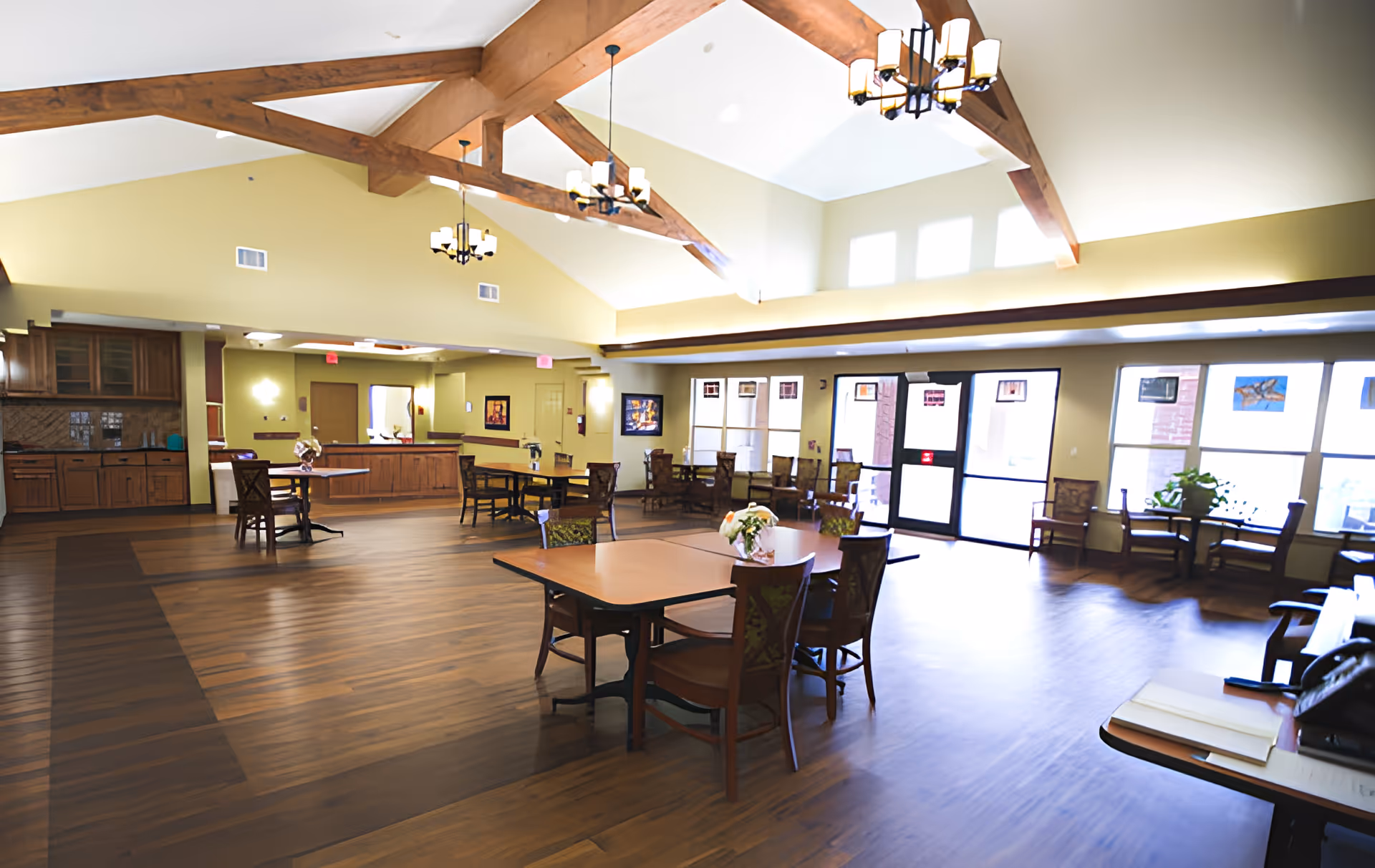 Spacious and well-lit common area with wooden floors, multiple tables and chairs, high vaulted ceiling with exposed wooden beams, chandeliers, and large windows letting in natural light. There is a kitchen area with wooden cabinets and a countertop in the background.