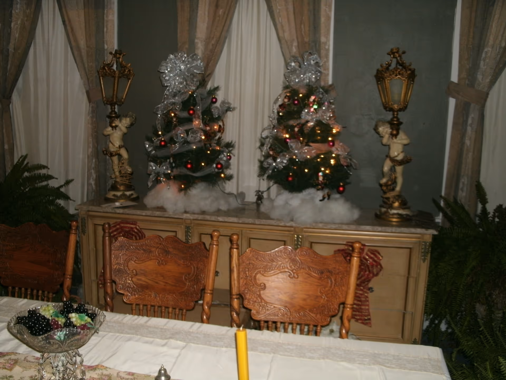 A dining room with a wooden table covered by a white tablecloth and ornate wooden chairs. On the table is a glass bowl filled with artificial grapes and a yellow candle. Behind the table is a sideboard with two small decorated Christmas trees adorned with silver ribbons and red ornaments, flanked by two vintage-style lamps held by cherub statues. The background features windows with sheer curtains and heavier drapes, and there are green plants on either side of the sideboard.