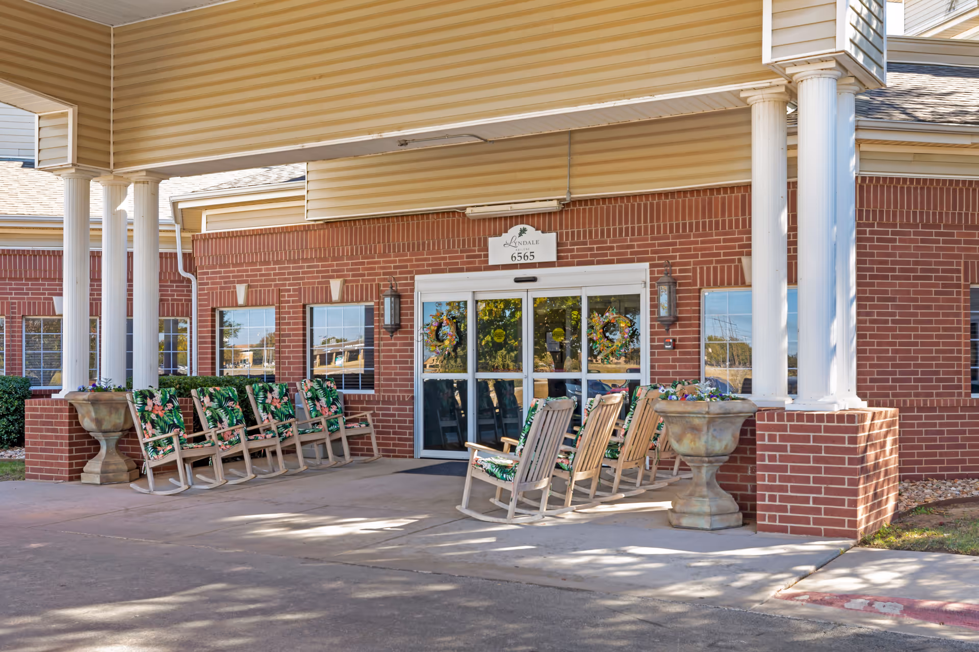 Entrance of Lyndale Abilene Senior Living facility showing a covered porch area with several wooden rocking chairs that have green floral cushions. The building has red brick walls, white columns, and glass double doors with decorative wreaths. Two large planters flank the entrance.