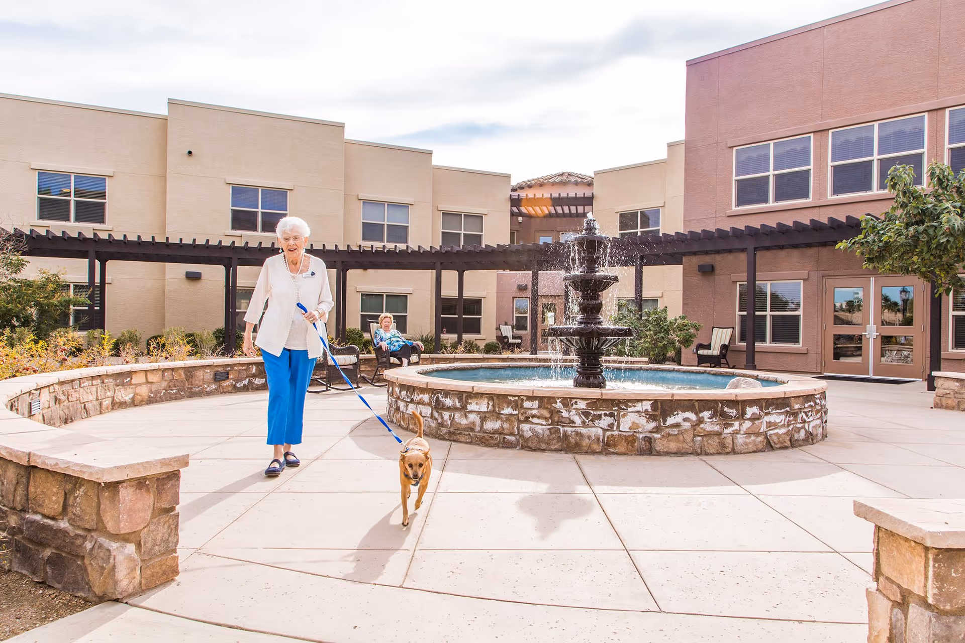 Elderly woman walks a dog in a sunny outdoor courtyard with a fountain.