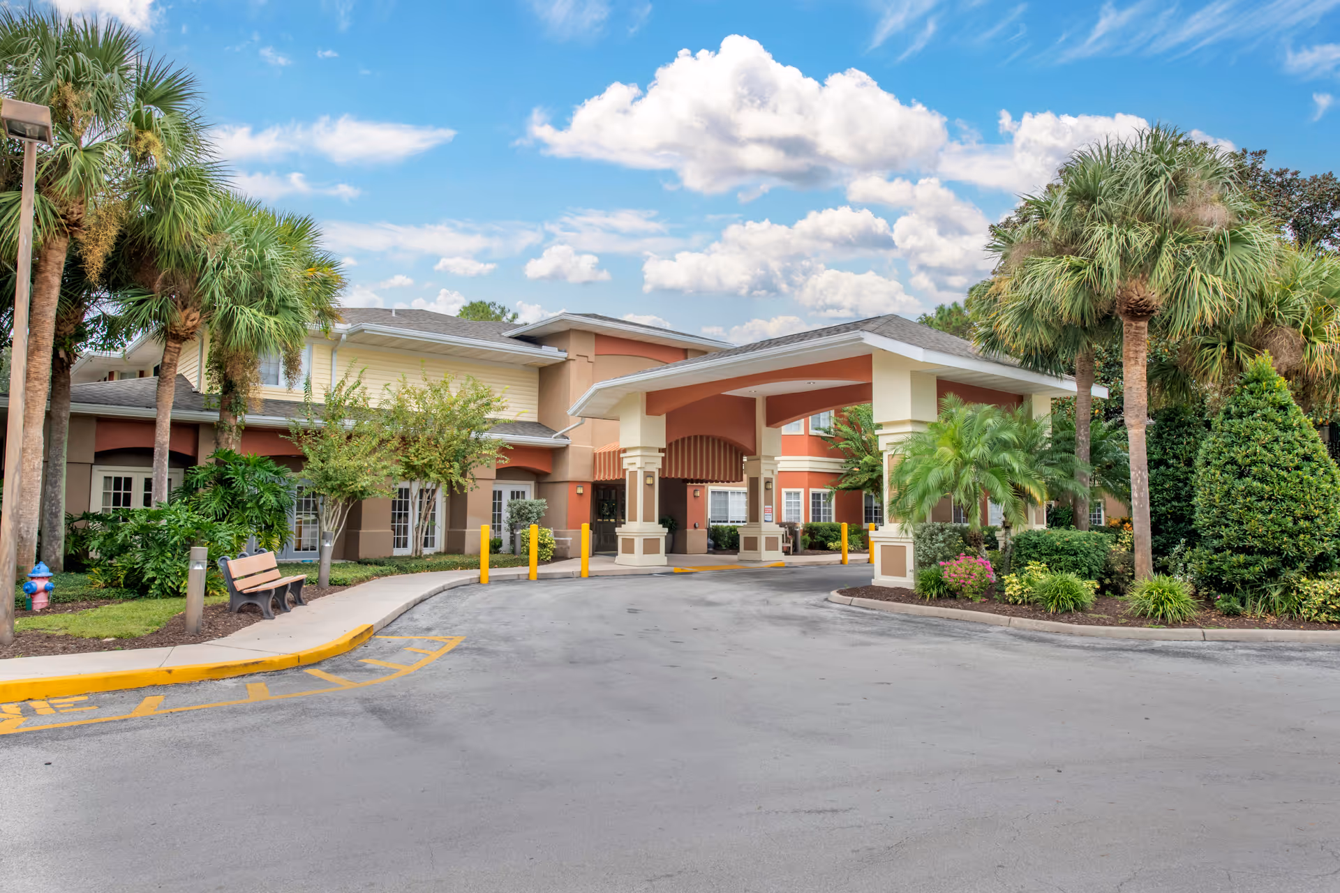 Exterior view of Brookdale Altamonte Springs senior living facility showing the entrance with a covered driveway, surrounded by palm trees and landscaped greenery under a partly cloudy blue sky.