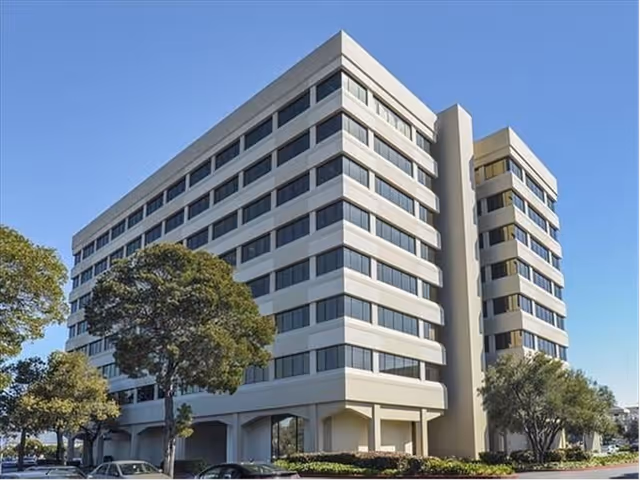 A tall, modern multi-story senior facility with trees lining the street and blue sky.