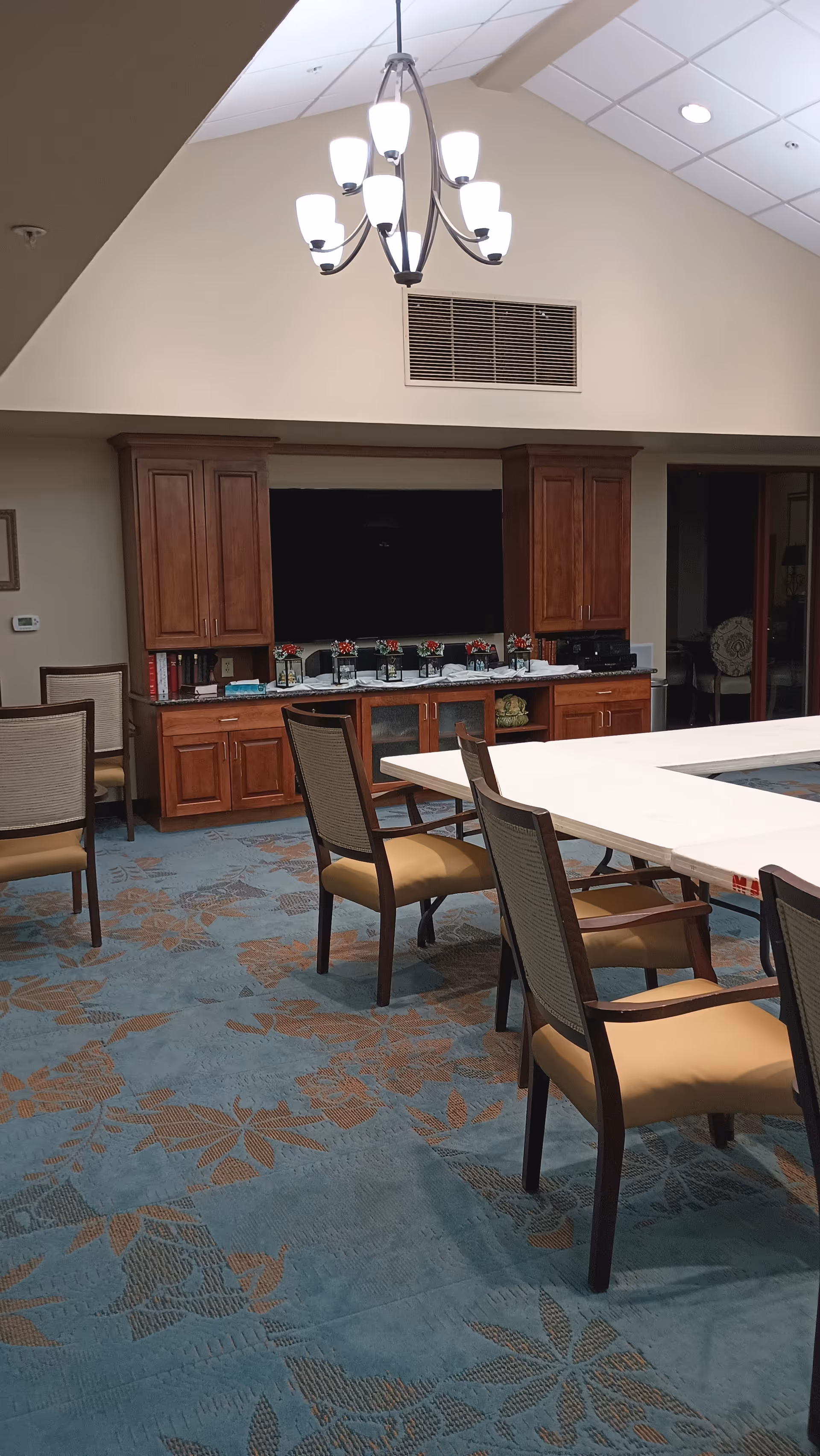 Spacious senior living dining area with a long table, chairs, chandelier, and dark wooden cabinets.