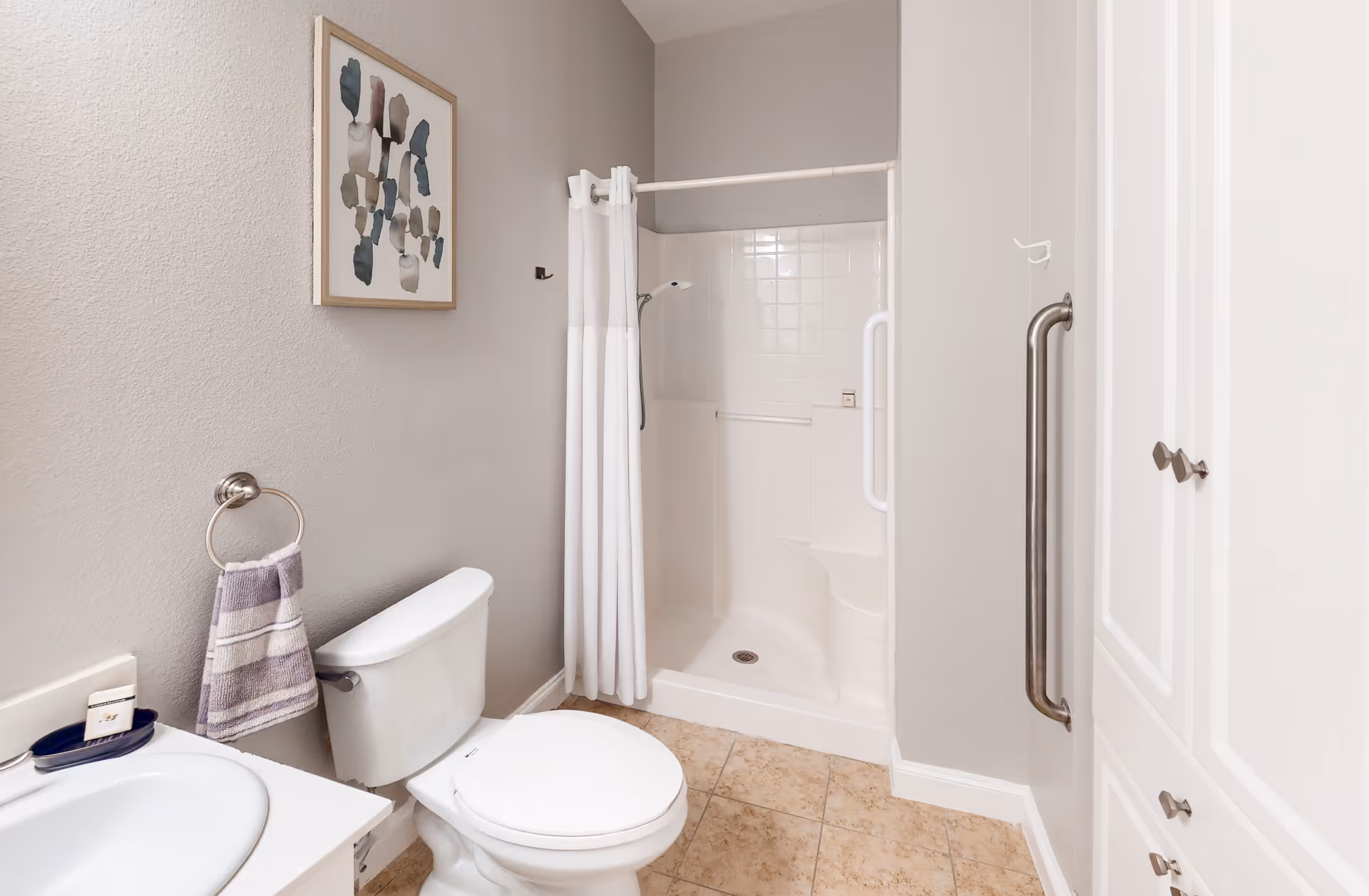 A clean bathroom featuring a white toilet, a sink with a soap dish, a towel ring with a striped hand towel, a walk-in shower with a white curtain and built-in seat, beige tiled floor, light gray walls, a large grab bar on the wall, and a framed abstract painting.