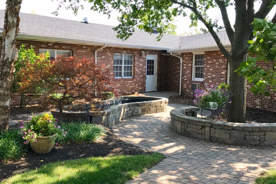 Brick single-story building with a paved courtyard, raised stone planters, trees, and potted flowers in front of a white door.