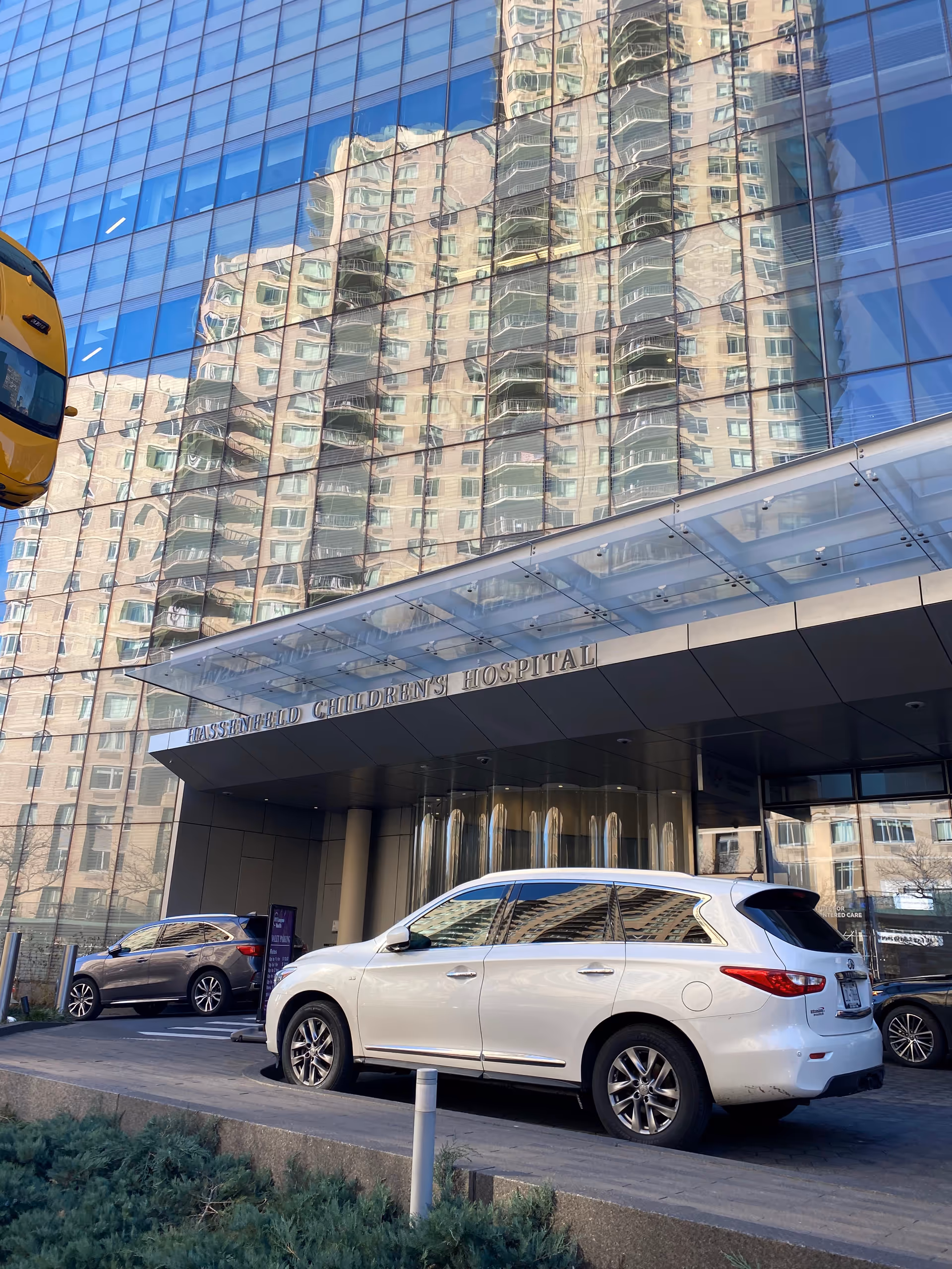 Glass-front hospital entrance with a reflection of a tall building and parked cars.