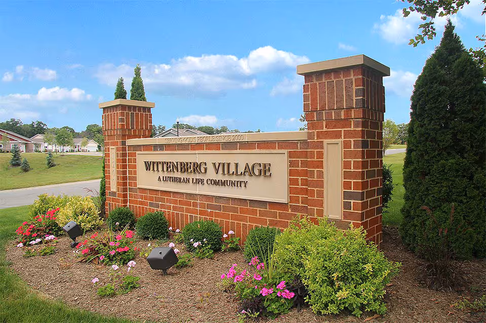 Brick entrance sign reading "Wittenberg Village, A Lutheran Life Community" set in landscaping with flowers, lawn, and houses in the background.