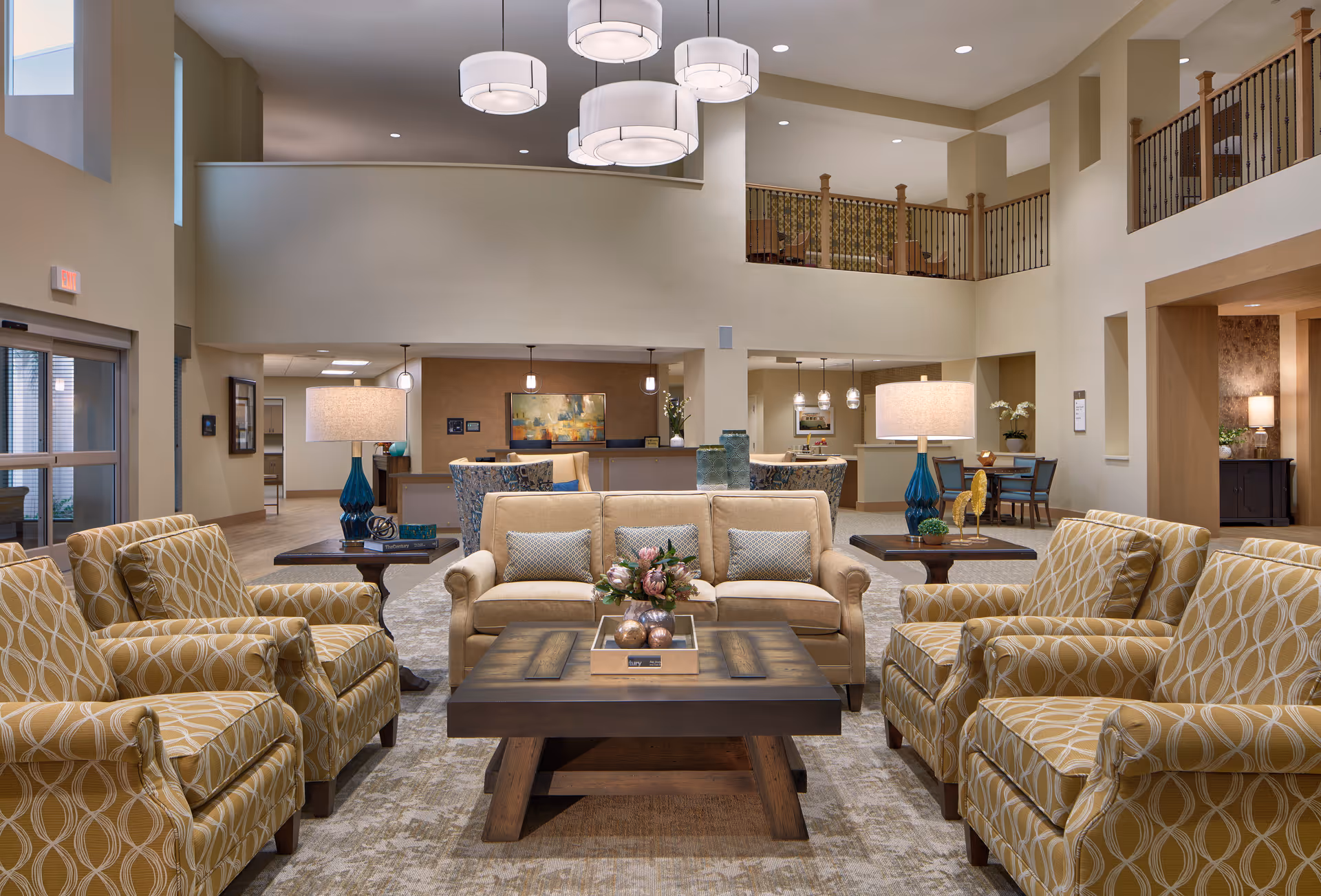 Spacious two-story senior living lobby with sofas, patterned armchairs, coffee tables, pendant lights, and a reception desk in the background.