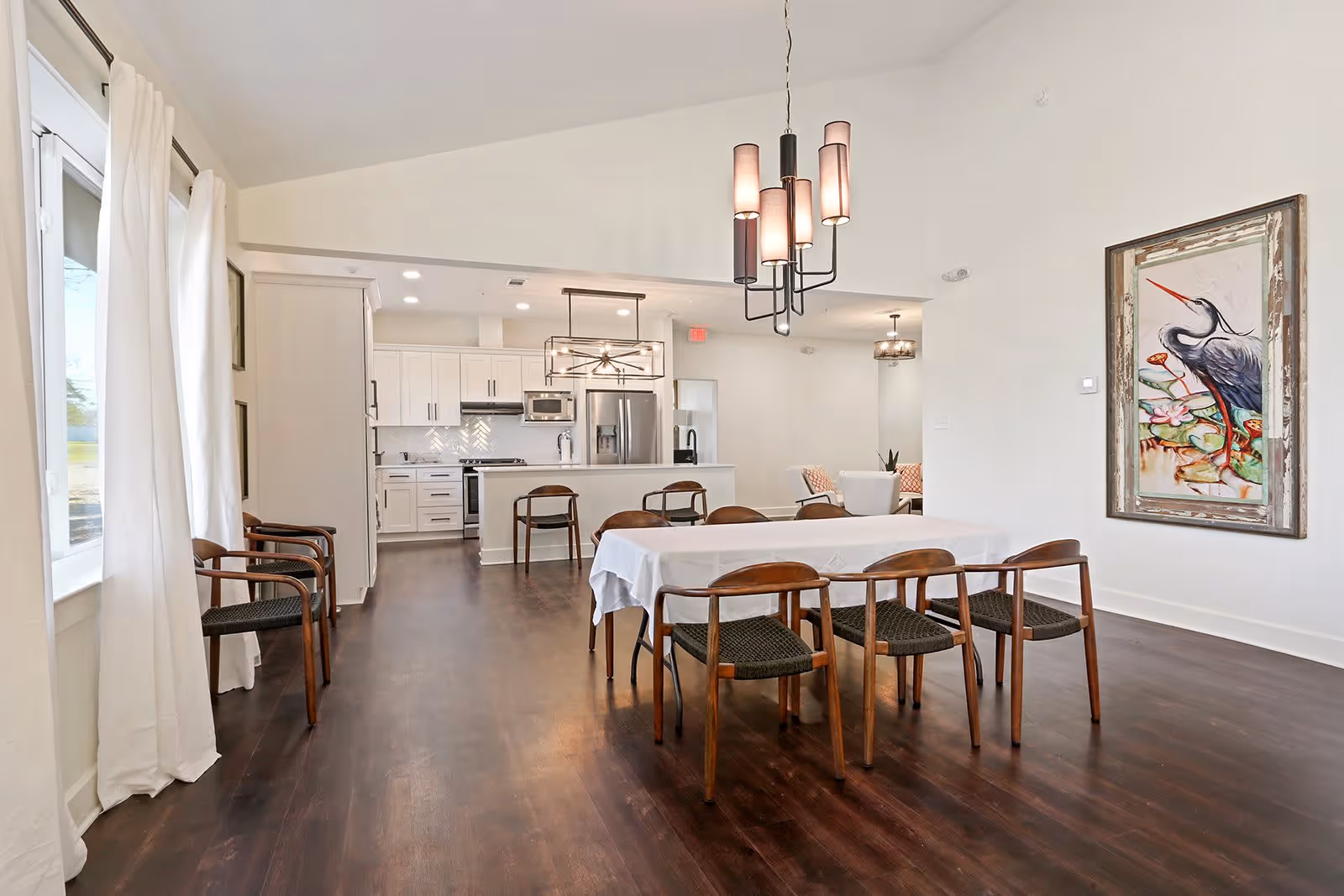 A spacious and modern dining area with a rectangular table covered with a white tablecloth surrounded by eight wooden chairs with woven seats. The room features dark hardwood flooring, white walls, and large windows with white curtains on the left side. In the background, there is a white kitchen with stainless steel appliances, including a refrigerator, microwave, and stove. Two contemporary pendant lights hang from the ceiling, one above the dining table and one above the kitchen island. A framed painting of a bird with a long beak standing among water lilies is mounted on the right wall.
