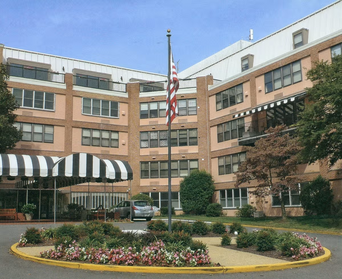Exterior view of a multi-story brick building with large windows, a circular driveway with a landscaped flower bed in the center, an American flag on a flagpole, and a black and white striped awning over the entrance.