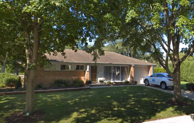 Single-story brick senior living facility front entrance with driveway and trees.