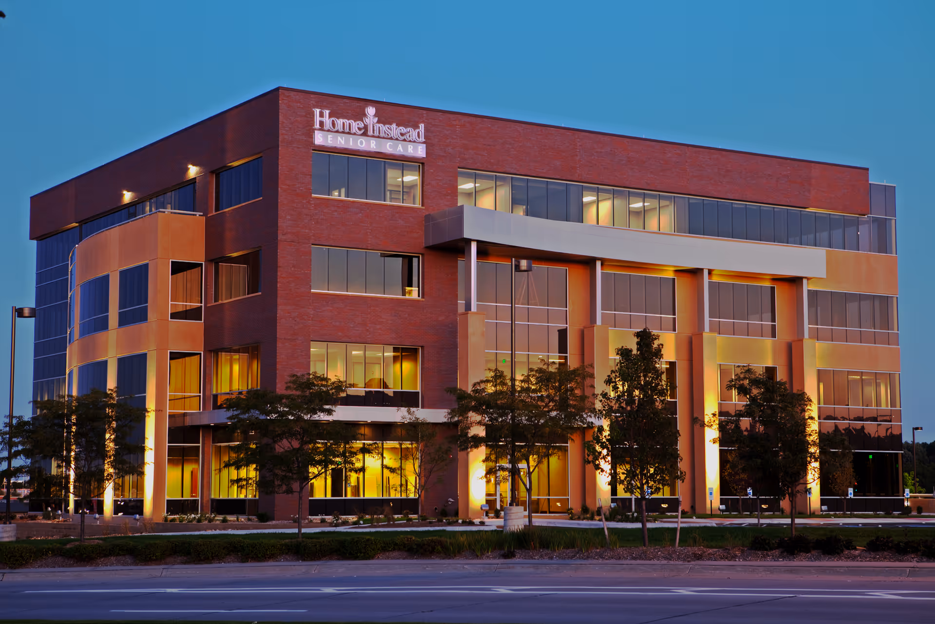 Modern brick office building with illuminated windows and a Home Instead Senior Care sign at dusk.