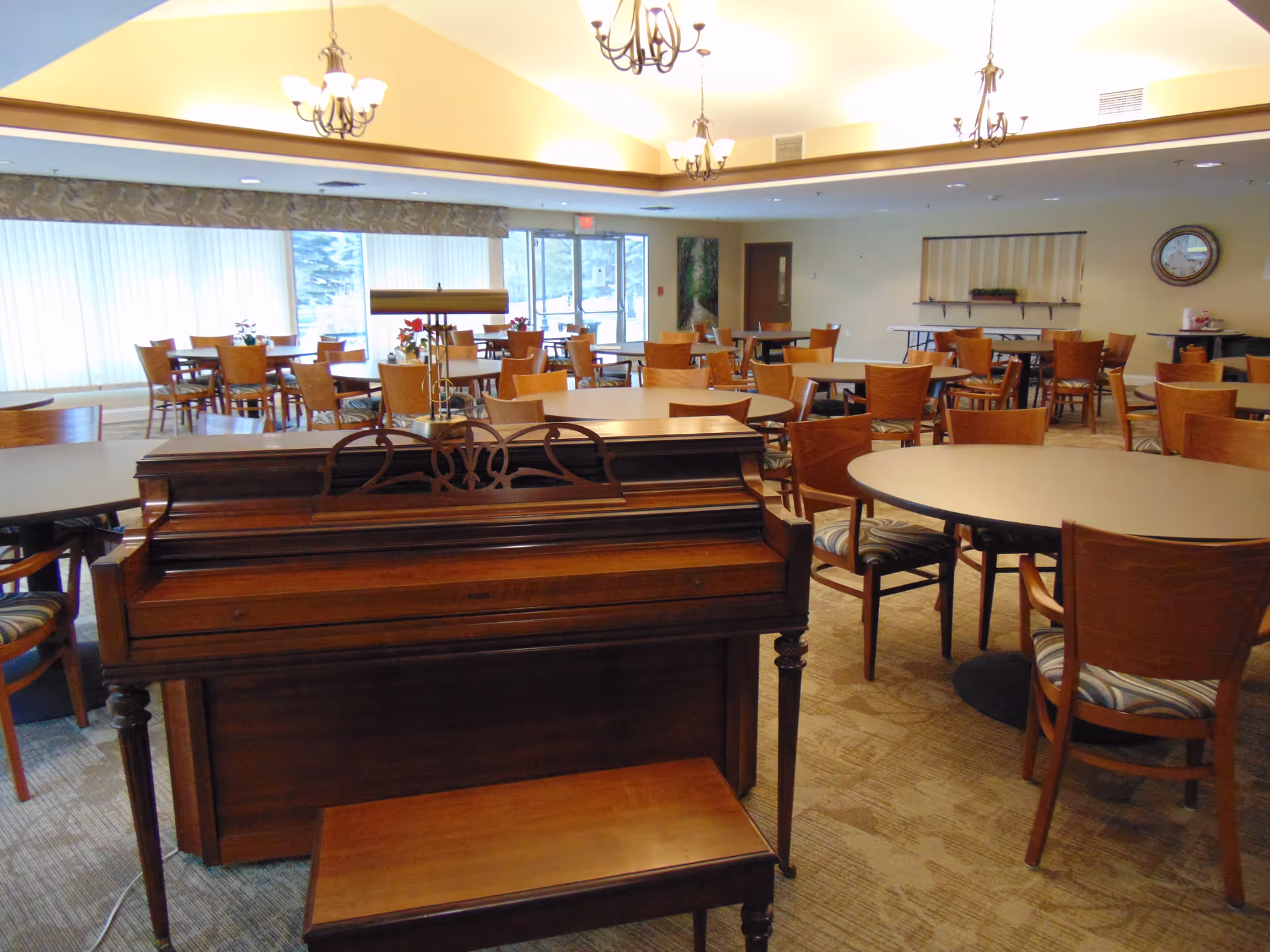 Spacious dining/activity room with a wooden piano in the foreground and multiple round tables and chairs beneath chandeliers.
