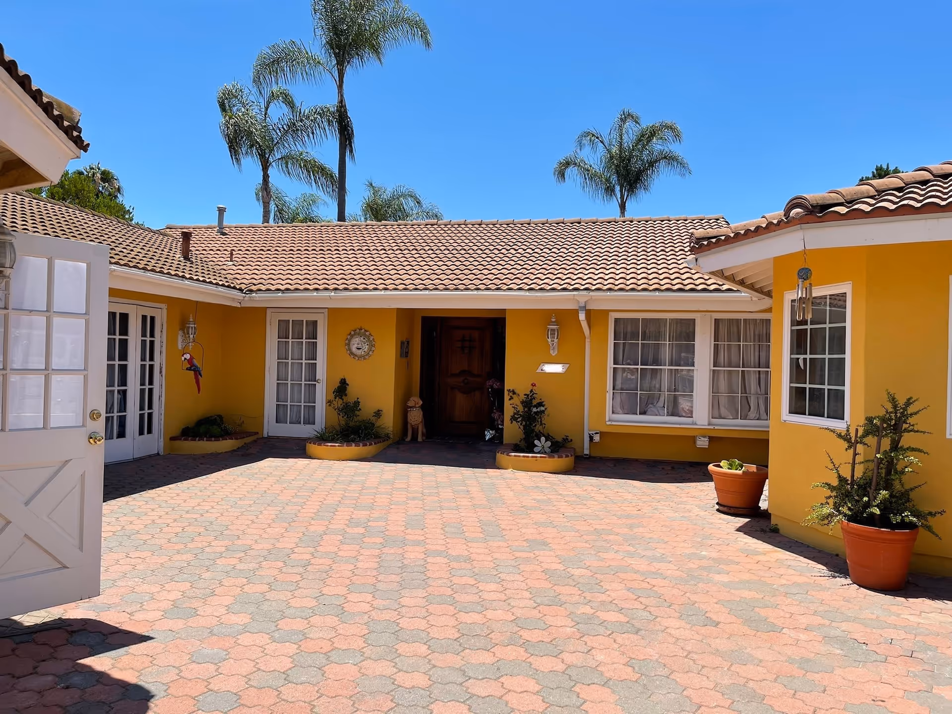 Exterior courtyard of a single-story building with yellow walls and a tiled roof under a clear blue sky. The courtyard is paved with patterned bricks and has several potted plants and small garden beds. There are multiple windows and doors, including a wooden front door in the center. Palm trees are visible in the background.