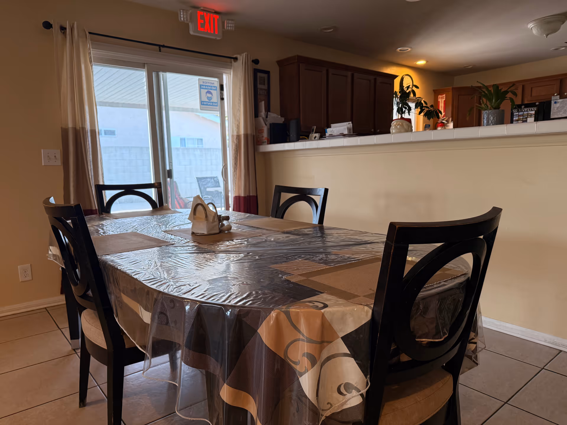 A dining room with a plastic table cover, chairs, sliding door, and a kitchen counter.