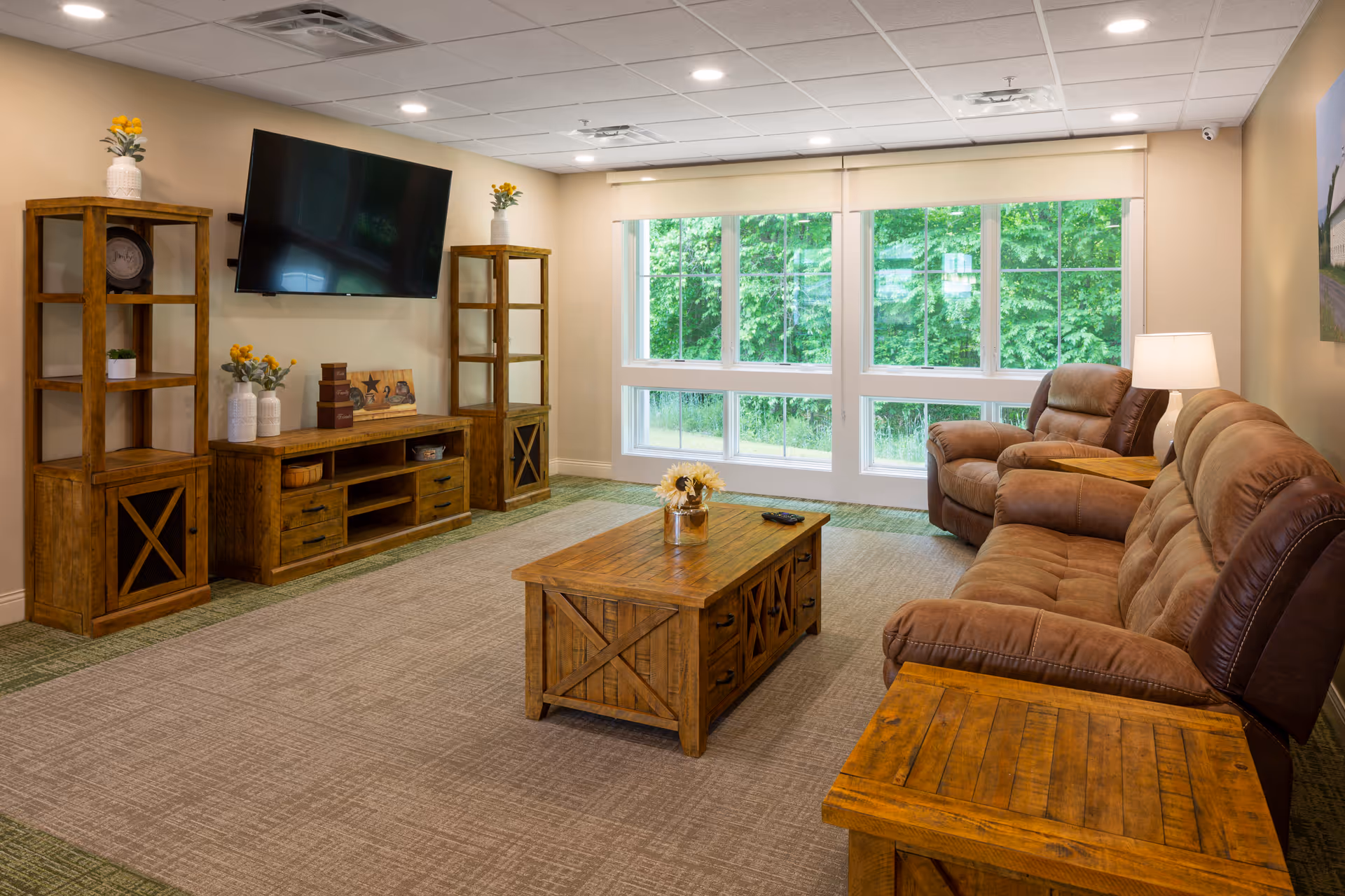 Cozy senior living lounge with brown leather seating, wooden tables, and a wall-mounted TV.
