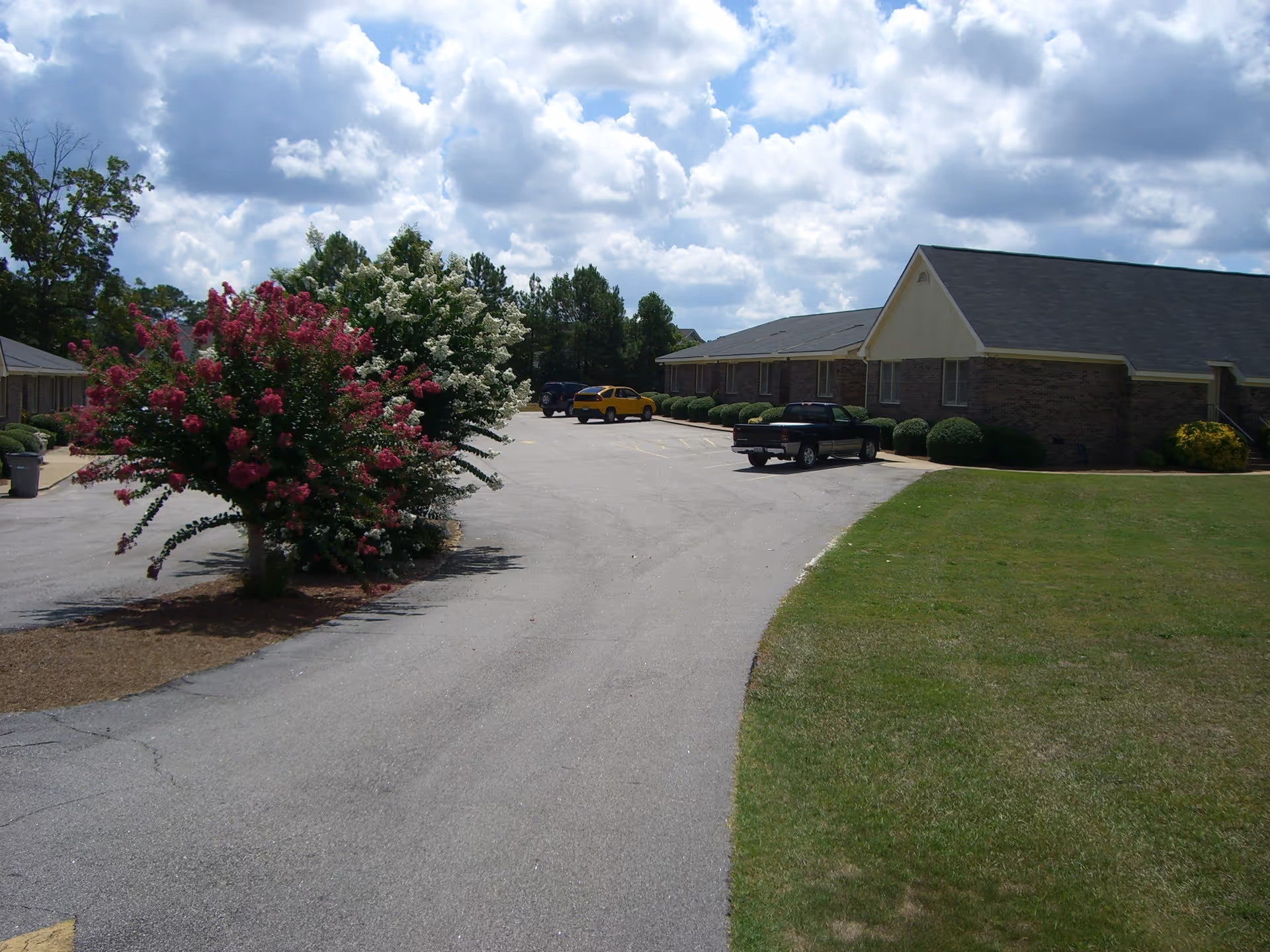 Parking lot area of a senior living facility with a few parked vehicles, surrounded by green grass and flowering trees under a partly cloudy sky. The building has a brick exterior with a dark shingled roof.
