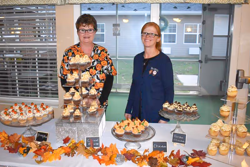 Two women standing behind a table with a variety of decorated cupcakes displayed on tiered trays and plates. The table is decorated with autumn leaves and small signs labeling the cupcake flavors such as Pumpkin, Caramel, Chocolate, and Heath Chocolate. The setting appears to be indoors with large windows and a door in the background.