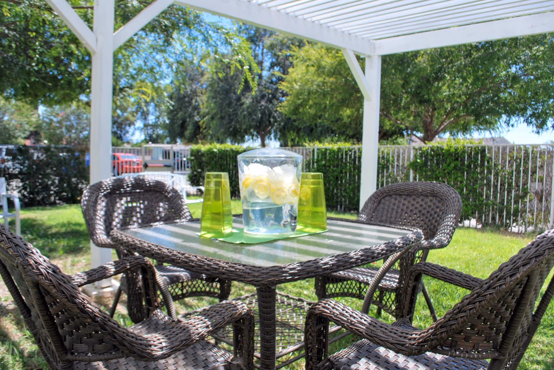 Wicker patio table and four chairs under a white pergola with a pitcher of lemon water and green glasses on the table in a sunny garden.