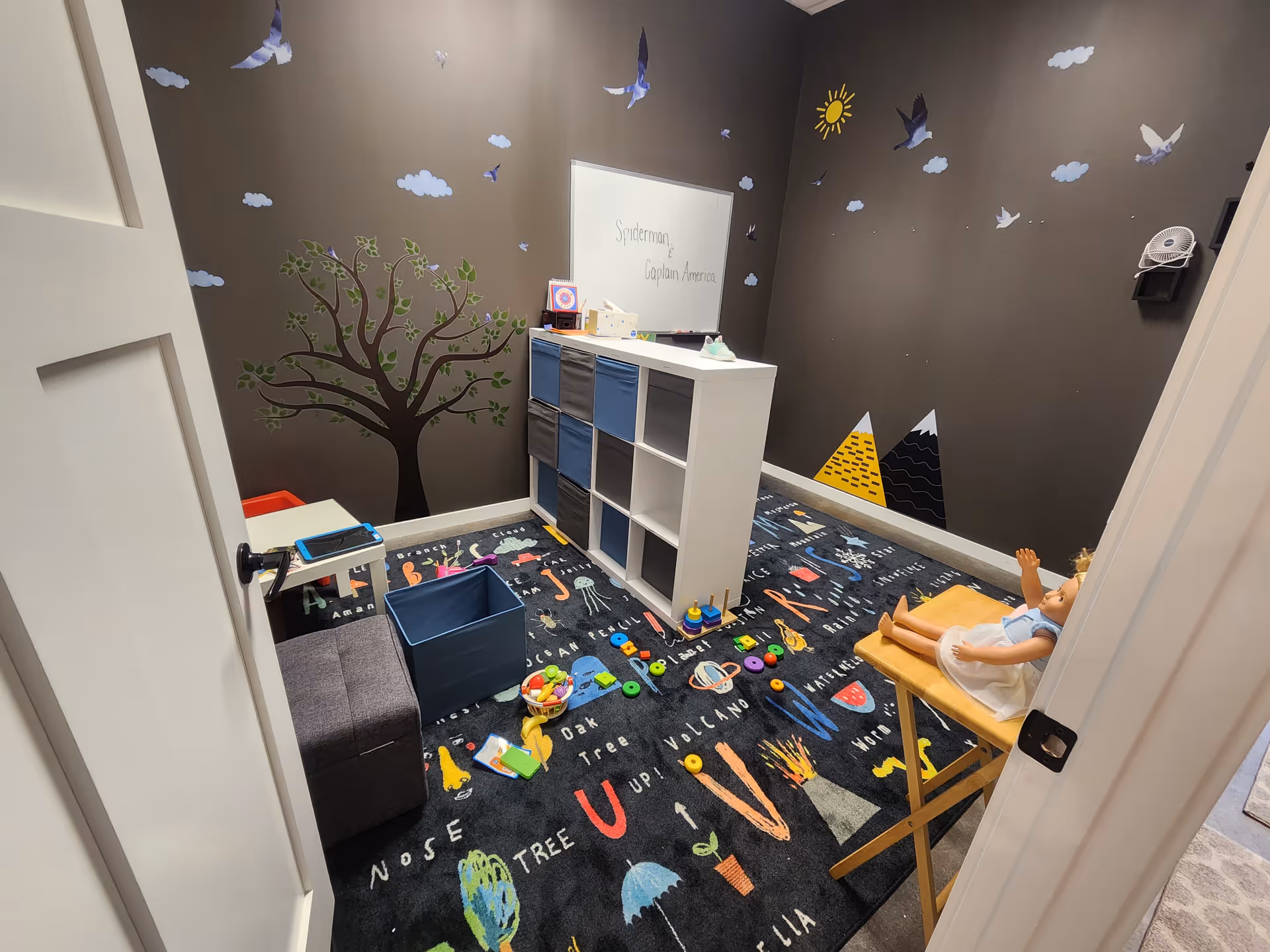 Play area with mural wall, cubby shelves, toys, and a therapy doll on a chair.