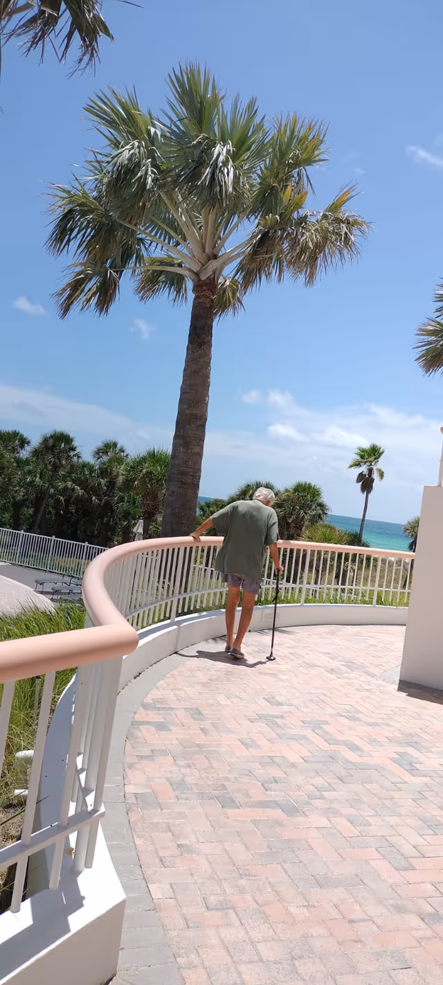 Senior with a cane walks along a curved outdoor walkway by palm trees.