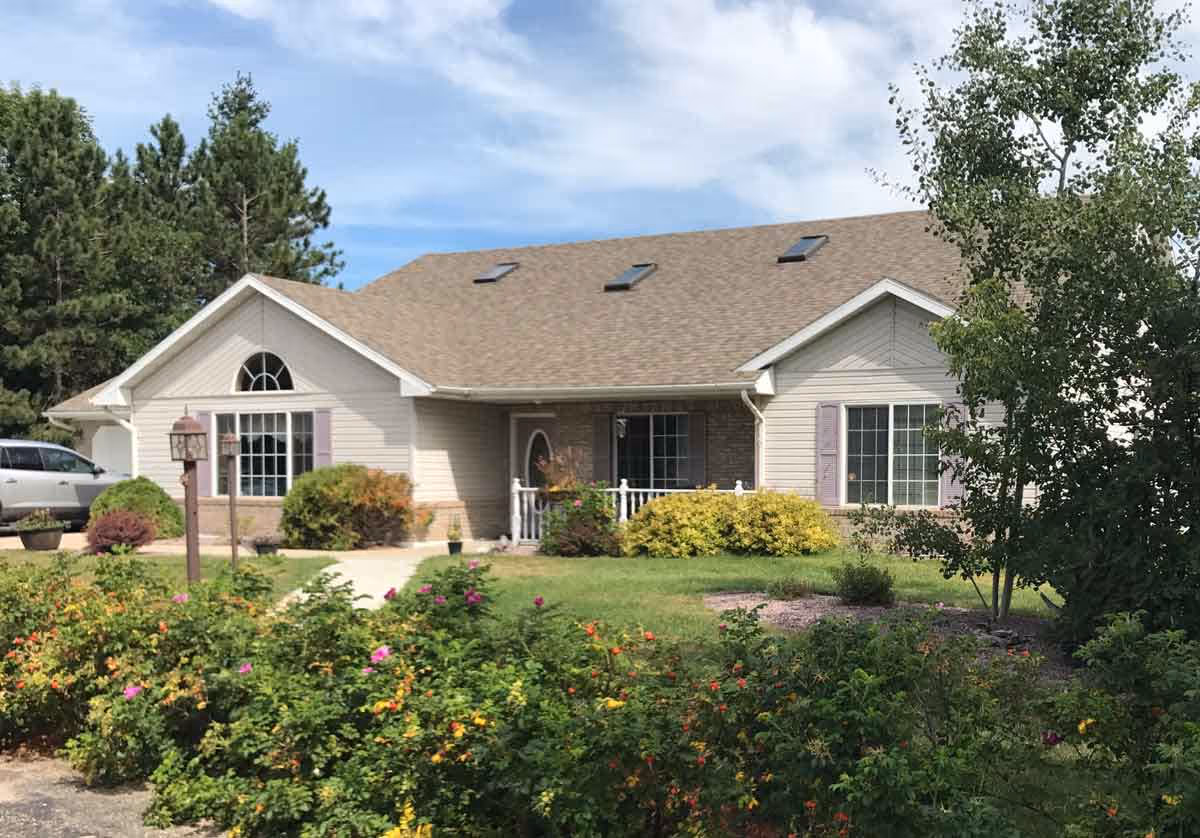 Single-story residential building with beige siding and a brown shingled roof, surrounded by green shrubs, flowering plants, and trees under a partly cloudy sky.