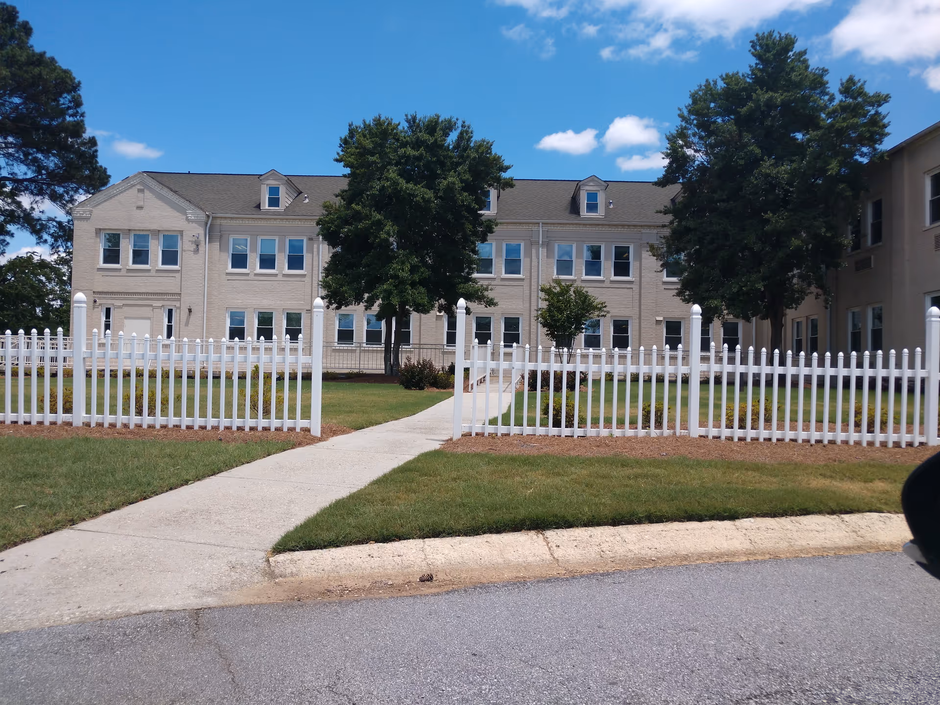Exterior view of a beige senior living building behind a white picket fence with trees.