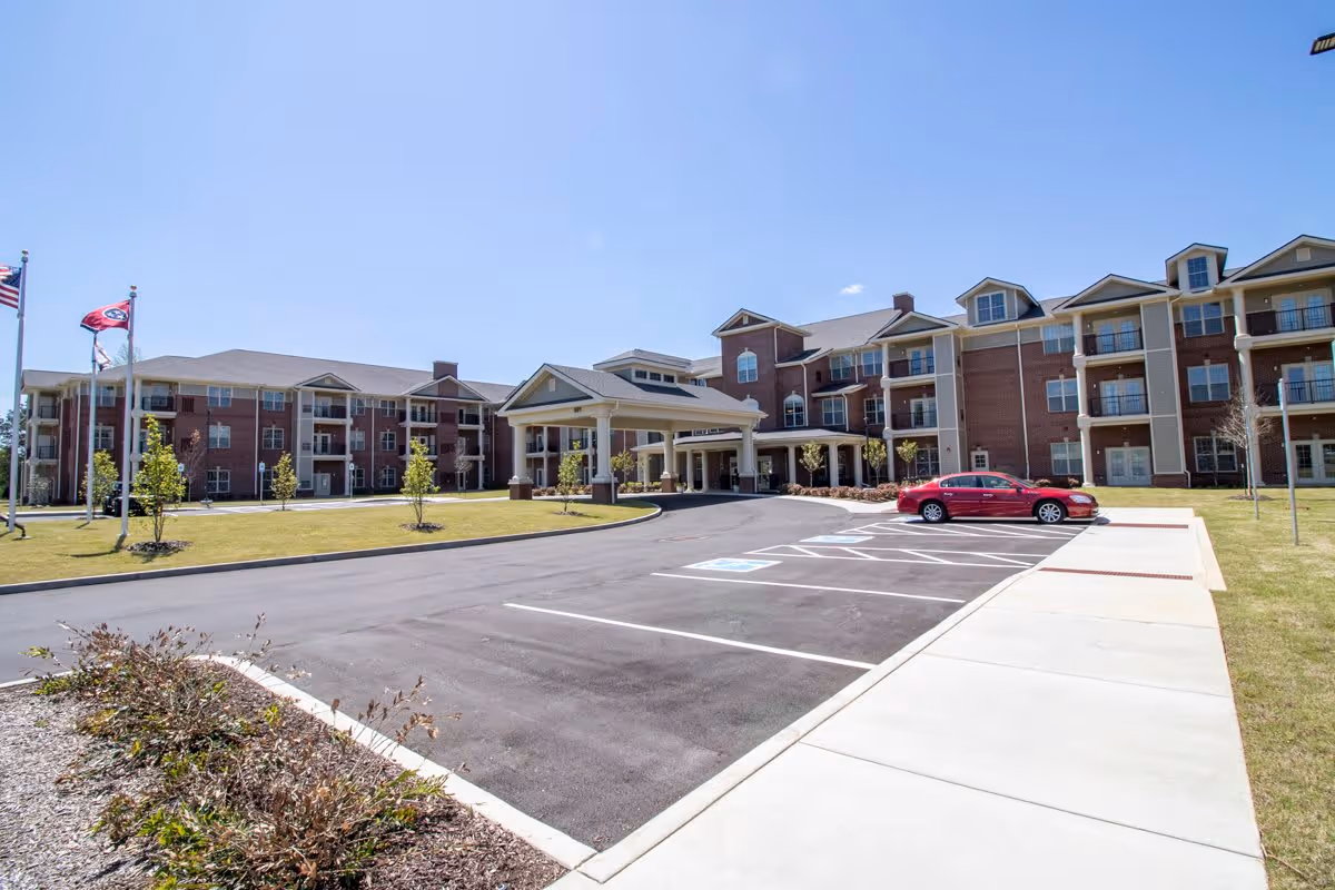 Exterior view of a large, multi-story senior living facility building with a covered entrance, several windows, balconies, and a parking lot with a red car parked. There are three flagpoles with flags on the left side and a clear blue sky above.
