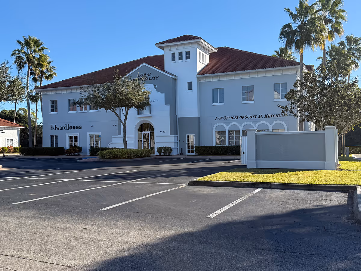 Front view of a light-gray office building with palm trees and a parking lot.