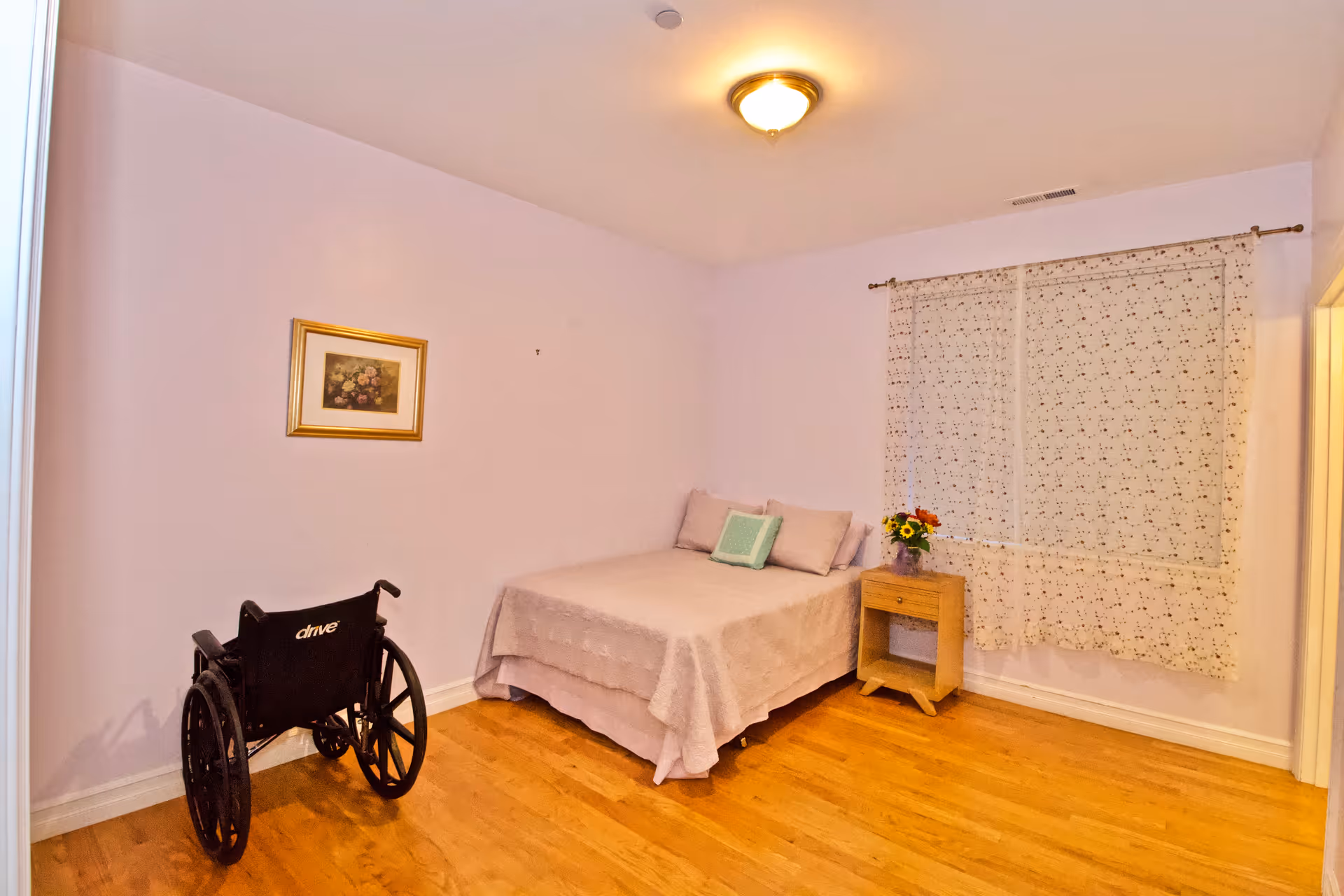 Simple bedroom in a senior facility featuring a bed, wheelchair, nightstand, and curtains.