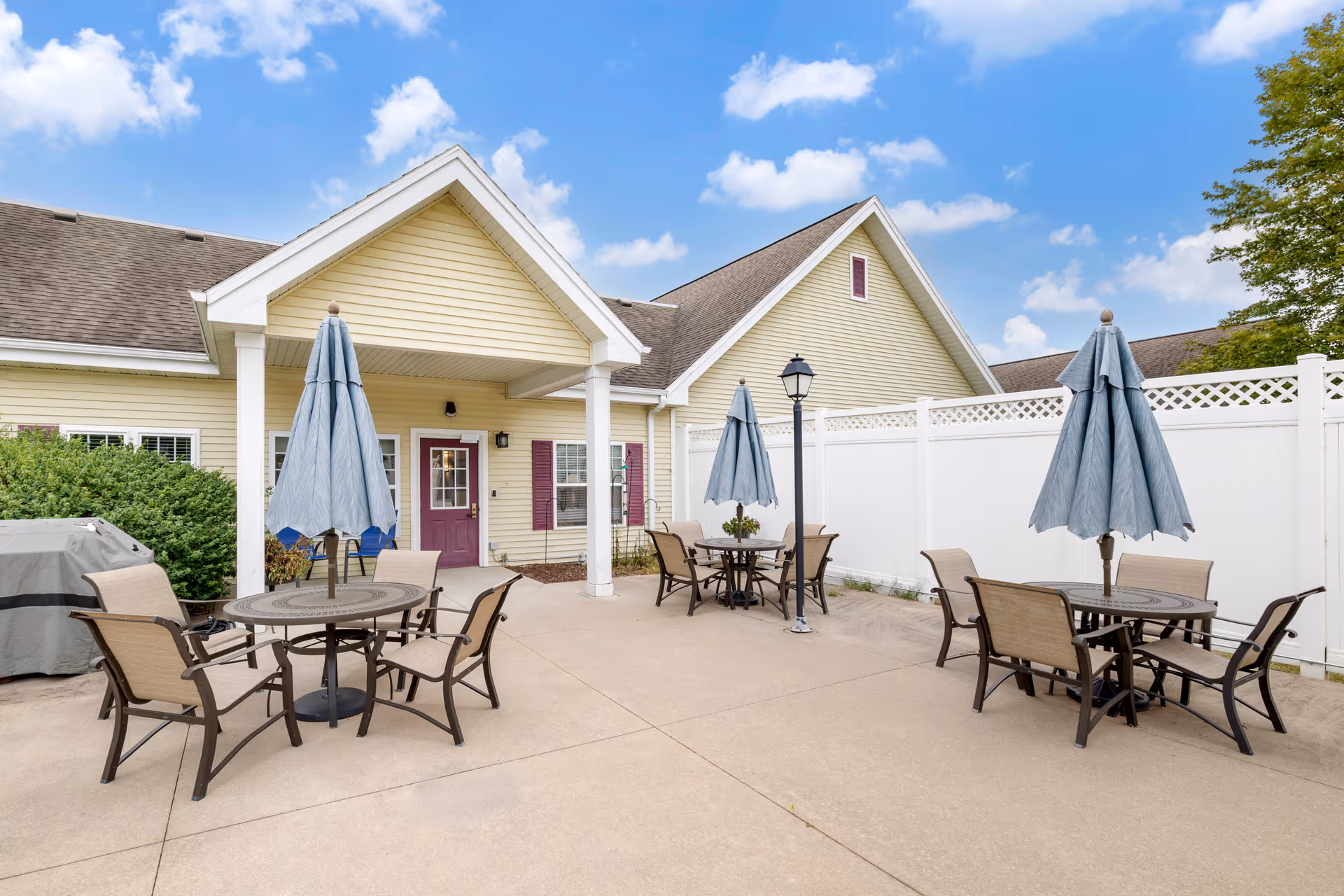 Outdoor patio at Noelitz Home with tables, chairs, and blue umbrellas beside a yellow building.