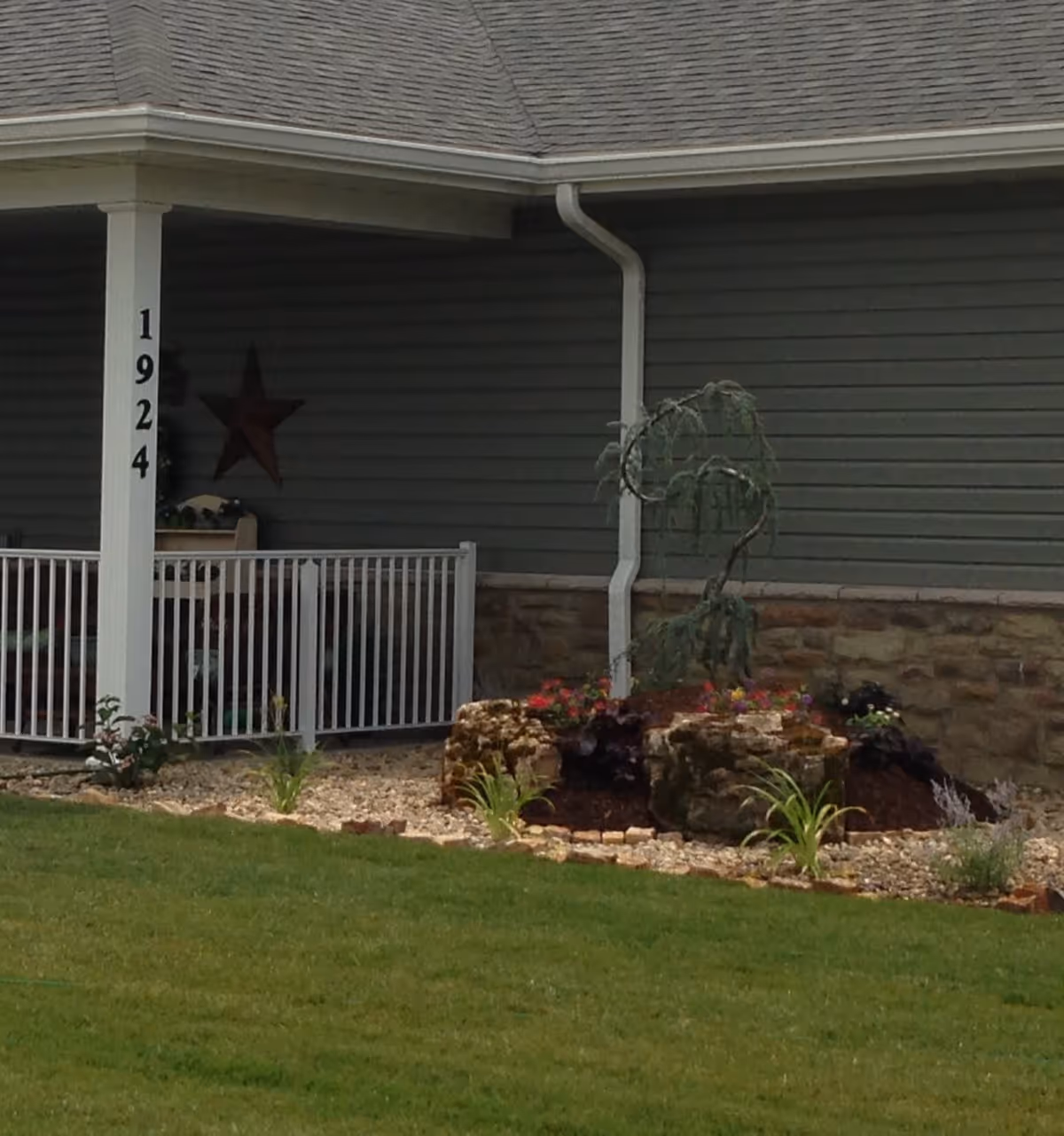A section of a building exterior with gray siding and a stone base. There is a covered porch area with a white railing and a white post displaying the number 1924. In front of the building is a landscaped garden bed with rocks, small plants, and flowers. A small tree with drooping branches is also visible in the garden bed. The lawn in the foreground is green and well-maintained.