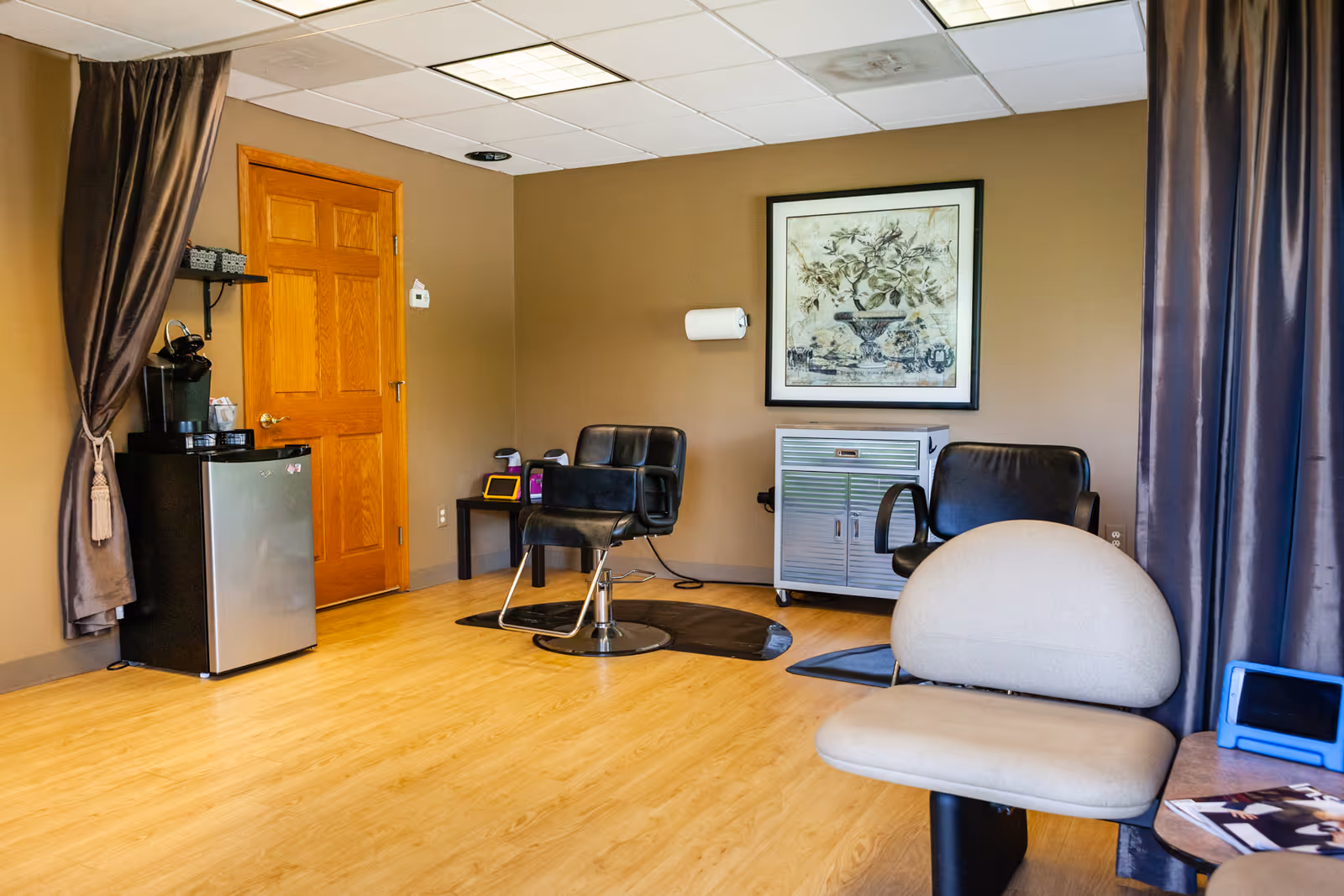 Interior salon area with styling chairs, a mini fridge, cabinet, and framed artwork.