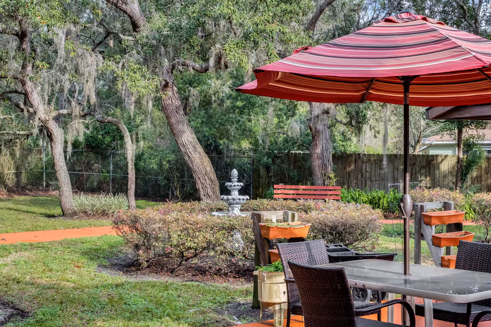 Outdoor garden area with a red and pink striped umbrella over a table and chairs. There are several planters with greenery, a red bench, a multi-tiered fountain, and large trees with hanging moss in the background.