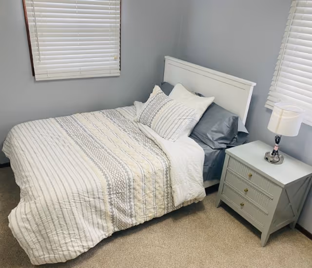 Neatly made bedroom in a senior living facility with a bed, striped quilt, pillows, and nightstand.