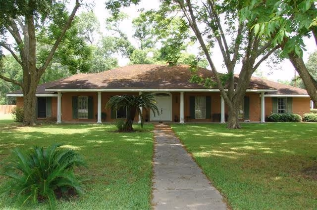 Front view of a single-story brick building with a covered porch, central walkway, lawn and large shade trees.