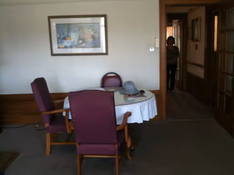 Small dining area with a round table covered by a white cloth, three purple chairs, a hat and magazines on the table, a framed picture on the wall and a person standing in a hallway doorway.